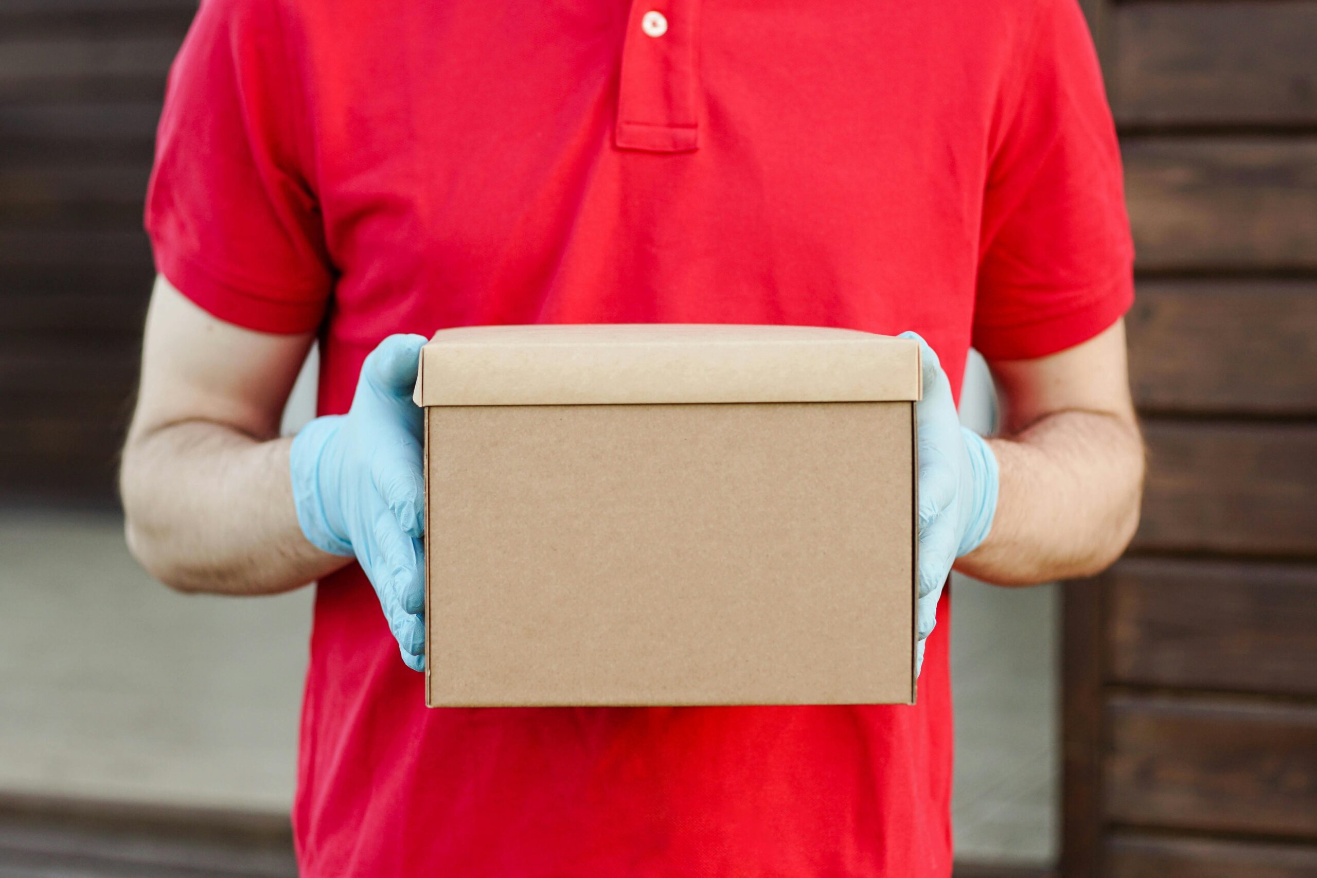 A delivery person in a red polo shirt and gloves holds a cardboard box, ready for delivery.